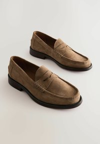 Mocasines de ante marrón con un diseño de mocasín, detalles de costura y una suela de cuero suave. Presenta una costura elevada sutil a lo largo del empeine.
