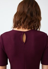 Bordeaux jurk met korte mouwen, heeft een sleutelgat-uitsnijding aan de achterkant en een textuurstof. Eenvoudige ronde halslijn.