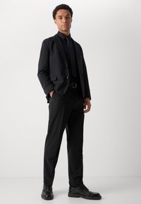 Black suit with a slim fit, single-button blazer and matching trousers. Worn with a black dress shirt and tie, complemented by black shoes.