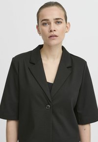 Juodas trumpas rankovėmis švarkas su atlenkiamu kraštu ir vienu mygtuku. Pagamintas iš struktūrizuotos medžiagos, turinčios lygią tekstūrą.