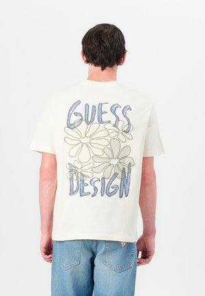 Kremfarget t-skjorte med blå "GUESS" og grønn "DESIGN" tekst, med blomsteromriss på baksiden. Korte ermer og avslappet passform.