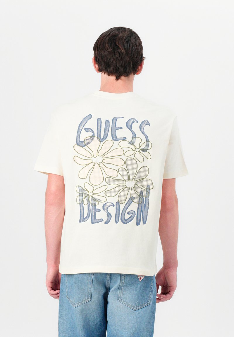 Crèmekleurig T-shirt met blauwe tekst "GUESS" en groene tekst "DESIGN", met bloemomtrekken op de rug. Korte mouwen en een casual pasvorm.