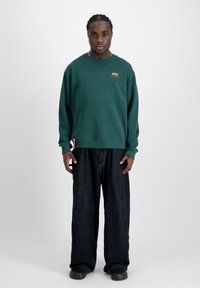 Felpa verde con scollo rotondo, polsini a coste e un piccolo logo giallo. Abbinata a jeans scuri a gamba larga e calzature nere.