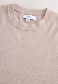 Ljusbeige ribbad tröja med rund halsringning, med en etikett märkt "NEXT" och subtil textur. Tyget verkar mjukt och töjbart.