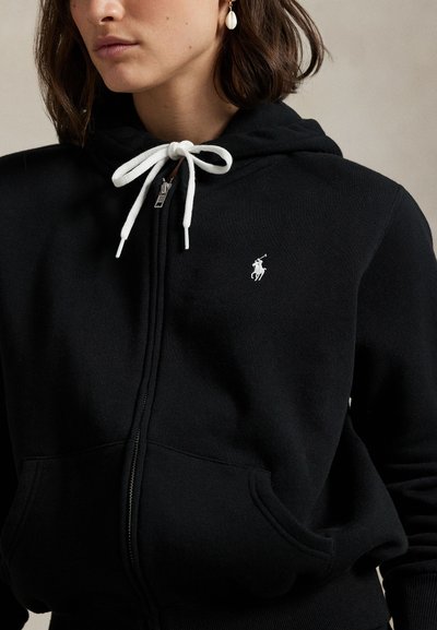 Žena v černé mikině na zip s bílými šňůrkami a malým bílým logem koně na levé straně hrudi, a zlaté náušnici s bílým přívěskem.