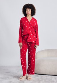 Κόκκινο σετ πιτζάμας με πουκάμισο με κουμπιά και παντελόνια με ελαστική μέση, και τα δύο με λευκά σχέδια καρδιάς. Μαλακό ύφασμα και άνετη εφαρμογή.