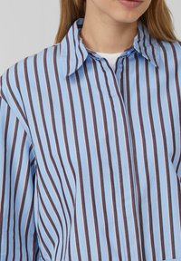 Camicia a righe blu e bianche con colletto a punta, caratterizzata da linee verticali e chiusura con bottoni. Tessuto in cotone leggero e liscio.