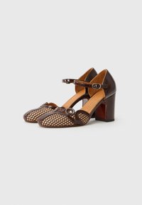 Scarpe con tacco marroni con una parte superiore in pelle verniciata e pannelli in rete con ritagli circolari. Il design include una cinghia con una fibbia rotonda.
