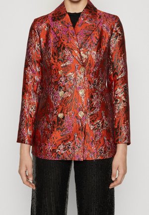 Blazer rojo de doble botonadura con patrones abstractos metálicos en dorado, rosa y negro, combinado con pantalones negros texturizados.