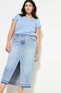 Camiseta azul claro con adornos en el hombro izquierdo, combinada con una falda de mezclilla de longitud media, ligeramente desgastada, que presenta una abertura en la parte delantera.