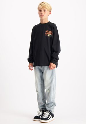 Schwarzes Langarmshirt mit gedrucktem Text und Grafik, kombiniert mit hellblauen, abgenutzten Jeans und schwarzen Sneakern mit weißen Streifen.