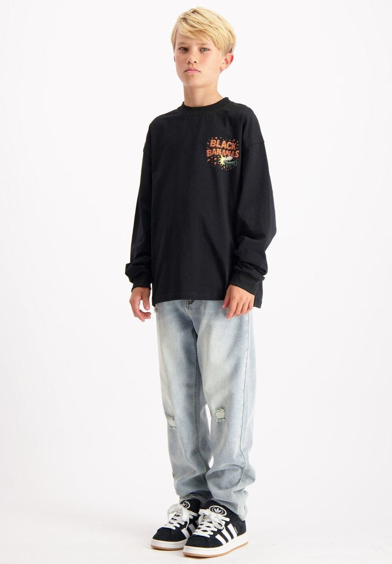 Zwart shirt met lange mouwen met bedrukte tekst en afbeelding, gecombineerd met lichtblauwe distressed jeans en zwarte sneakers met witte strepen.