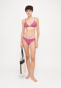 Bikini de estampado floral rosa com parte de cima triangular e cuecas correspondentes, confeccionado em tecido suave. Modelo a segurar uma mala tote preta e creme.