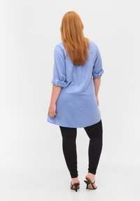 Vaaleansininen tunika, jossa on käärityt hihansuut, yhdistettynä mustiin legginsseihin ja korkokenkiin, näkyy takaa valkoista taustaa vasten.