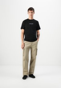 Jonge man staat gekleed in een zwart "Santa Cruz" t-shirt, beige cargobroek met zakken en zwarte sneakers tegen een effen witte achtergrond.