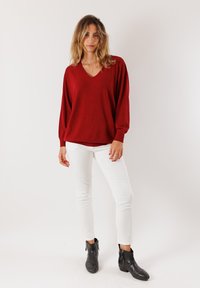 Maglione rosso oversize con scollo a V, maniche lunghe e polsini a coste. Abbinato a pantaloni bianchi slim-fit e stivali neri alla caviglia.