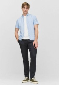 Lichtblauw overhemd met korte mouwen, wit T-shirt eronder, donkere broek en olijfgroene sneakers. Eenvoudig ontwerp, casual pasvorm en minimale hardware.