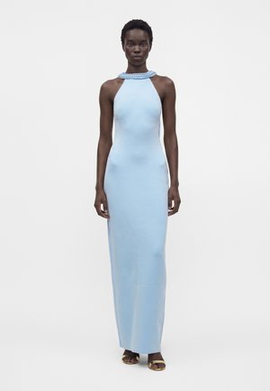Mujer con un vestido largo sin mangas de color azul claro con escote trenzado, de pie erguida con sandalias doradas sobre un fondo blanco.