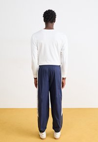 Camicia bianca a maniche lunghe e pantaloni da allenamento blu navy con accenti gialli, dotati di una vita elastica e strisce laterali. Sneakers bianche visibili.