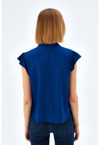 Marineblauwe mouwloze blouse met gerimpeld schouderdetail. Soepele textuur, licht gebogen zoom, in combinatie met donkere blauwe aansluitende jeans.
