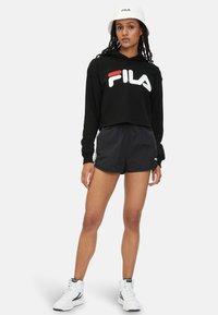 Sudadera corta negra con un atrevido logo "FILA" en blanco y rojo, combinada con pantalones cortos negros. Sombrero de cubo blanco y zapatillas altas blancas.