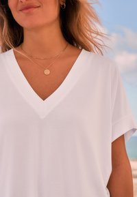 Chemise blanche à col en V avec manches courtes, en tissu lisse. Colliers superposés en or avec pendentif circulaire visible.