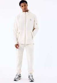 Man staat in een crèmekleurige hoodie en bijpassende broek, met wit en zwarte sneakers tegen een eenvoudige witte achtergrond.