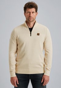 Lichtbeige ritstrui met een textuurpatroon, ribgebreide boorden en zoom, met een klein logopatch op de borst. Draag over een zwarte top.