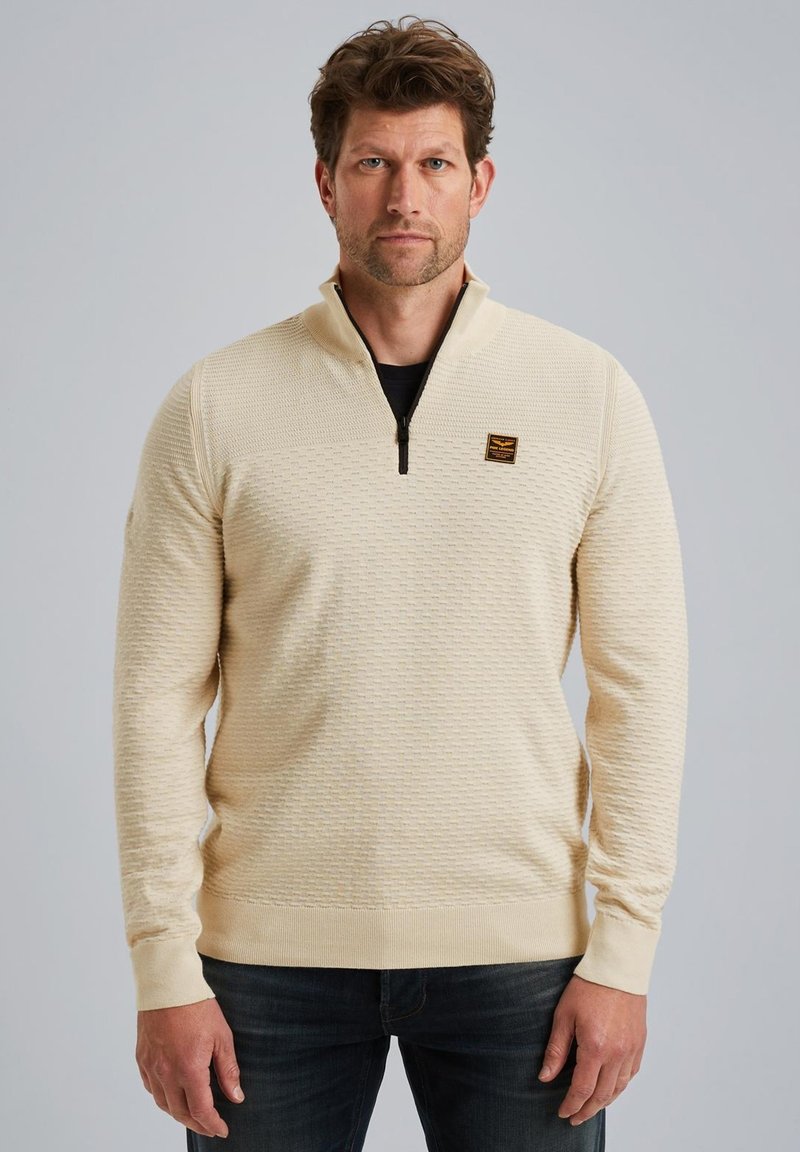 Lichtbeige ritstrui met een textuurpatroon, ribgebreide boorden en zoom, met een klein logopatch op de borst. Draag over een zwarte top.