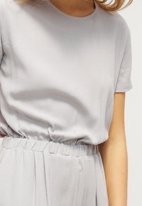 Jasnoszara bluzka o krótkich rękawach, elastycznym pasie oraz miękkiej teksturze materiału, charakteryzująca się luźnym krojem i gładkim układaniem.