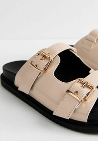 Sandalias de charol beige con dobles tiras y hebillas de hardware dorado, que cuentan con una suela negra texturizada y diseño de punta abierta.