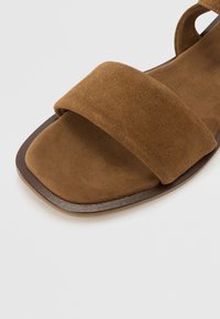 Bruna mockaskor med en bred rem som täcker fotryggen, platt sula och fyrkantig tådesign. Texturen är mjuk och slät.