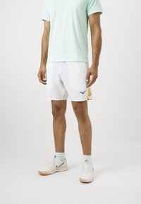 Hellgr�ünes Kurzarmhemd, weiße Shorts mit Navy-Logo und weiße Sportschuhe. Die Shorts sind leicht und haben eine glatte Textur.