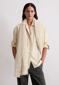 Vrouw met lang donker haar draagt een crèmekleurig overhemd met opgerolde mouwen, een beige sjaal met structuur en donker gestreepte broek, staande tegen een witte muur.