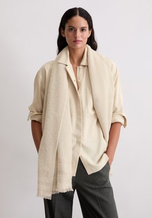 Femme aux longs cheveux sombres portant une chemise crème à boutons avec manches retroussées, une écharpe beige texturée et un pantalon à fines rayures sombre, debout contre un mur blanc.