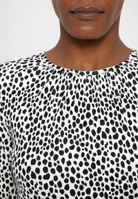 Gros plan d'une personne portant un haut blanc à pois noirs irréguliers et un col rond, montrant le bas du visage et le cou.