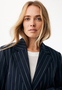 Granatowa marynarka w prążki wykonana z teksturowanej tkaniny; posiada ostre klapy i dopasowany krój, nałożona na biały dzianinowy top.