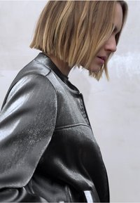 Metallinhohtoinen harmaa vetoketjullinen takki, jossa on sileä pinta; siinä on pystykaulus eikä näkyviä taskuja. Malli esitetään sivuprofiilissa.