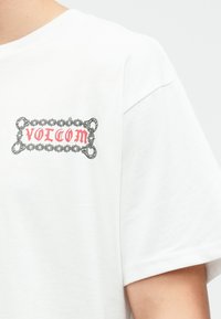 Witte katoenen t-shirt met een gedrukt logo in rood en zwart binnen een kettingontwerp, weergegeven op de linkerborst.