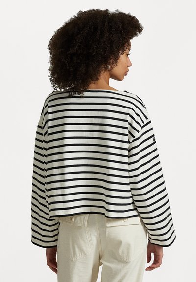 Haut à manches longues rayé en coton noir et blanc, avec un large col et des coutures d'épaule abaissées. Porté avec un pantalon de couleur claire.