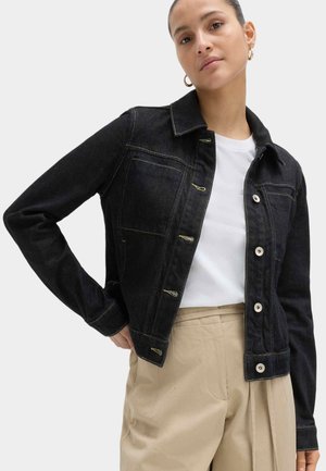 Kobieta pozująca z prawą ręką na biodrze, ubrana w czarną kurtkę jeansową, białą koszulę, beżowe spodnie oraz złote kolczyki koła na białym tle.