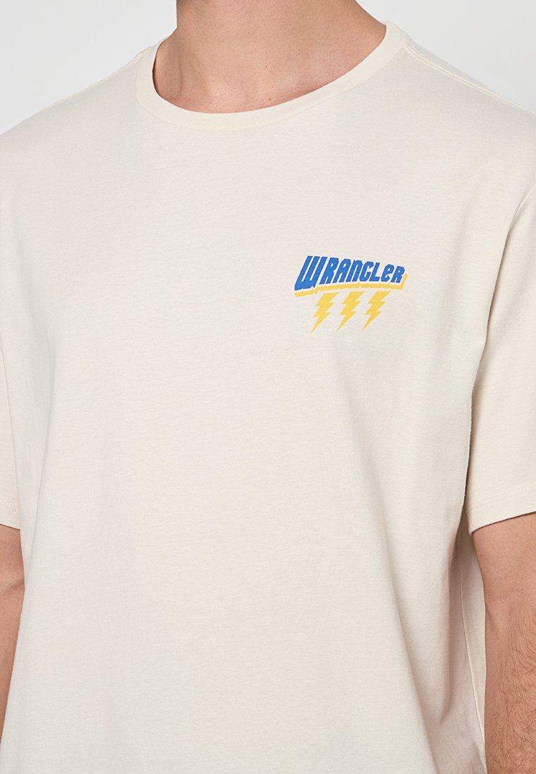 Camiseta de manga corta color crema hecha de algodón, que presenta un logo "WRANGLER" en azul y amarillo con rayos en el pecho.