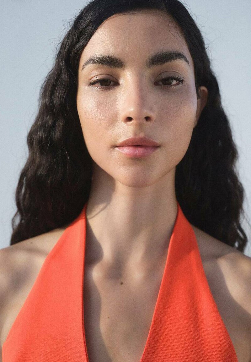 Close-up of a person with long, wavy black hair, wearing a sleeveless, deep V-neck orange top against a light background.