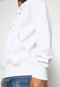 Person trägt einen weißen Calvin Klein Jeans Hoodie mit Fronttasche und blauen Jeans, seitlich auf Taillenhöhe gezeigt.