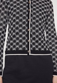 Cardigan a righe in bianco e nero con un design geometrico, bottoni dorati e rifiniture bianche lungo i bordi e le maniche.