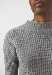 Maglione grigio lavorato a maglia con texture a coste, caratterizzato da un collo alto e maniche lunghe. Vista ravvicinata che mostra il dettaglio del tessuto.