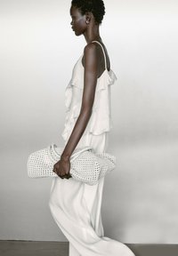 Helles graues, geschichtetes Rüschenshirt und fließende weiße Hose. Hält eine gewebte weiße Clutch-Tasche mit strukturiertem Oberflächen.