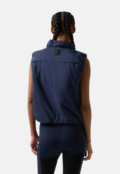 Kvinde med fletninger iført en ærmeløs marineblå dunvest og marineblå leggings, set bagfra på en ensfarvet lys grå baggrund.