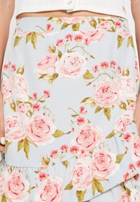 Jupe fleurie en tissu bleu clair ornée de grandes fleurs roses et de petites fleurs rouges, avec des feuilles vertes. L'ourlet à volant ajoute de la texture.