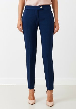 Kobieta ubrana w granatowe spodnie o dopasowanym kroju z przednią fałdą, zapinane na guzik i zamek błyskawiczny, zestawione z beżowymi szpilkami o spiczastym nosku.